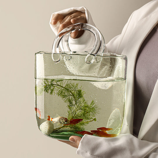 Clear handbag with water and small plants and fish, held by a person wearing a white shirt.