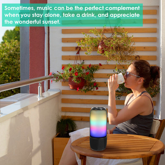 Woman sitting on a balcony with a colorful smart speaker, enjoying a drink and the sunset.