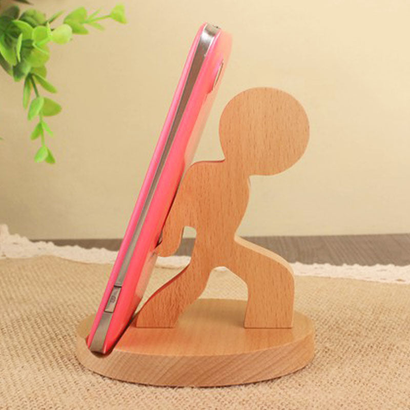 Wooden phone stand holding a pink phone on a beige surface with a plant in the corner.
