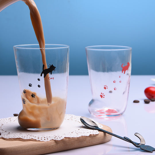 Glass with coffee being poured, featuring cat illustrations, on a table with another glass and coffee beans.