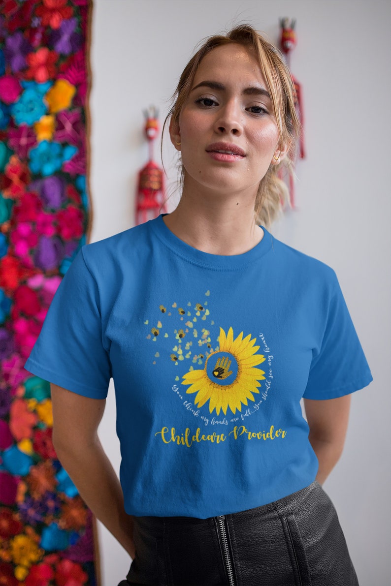 Woman wearing a blue t-shirt with a sunflower design and text, standing in front of a colorful floral wall.