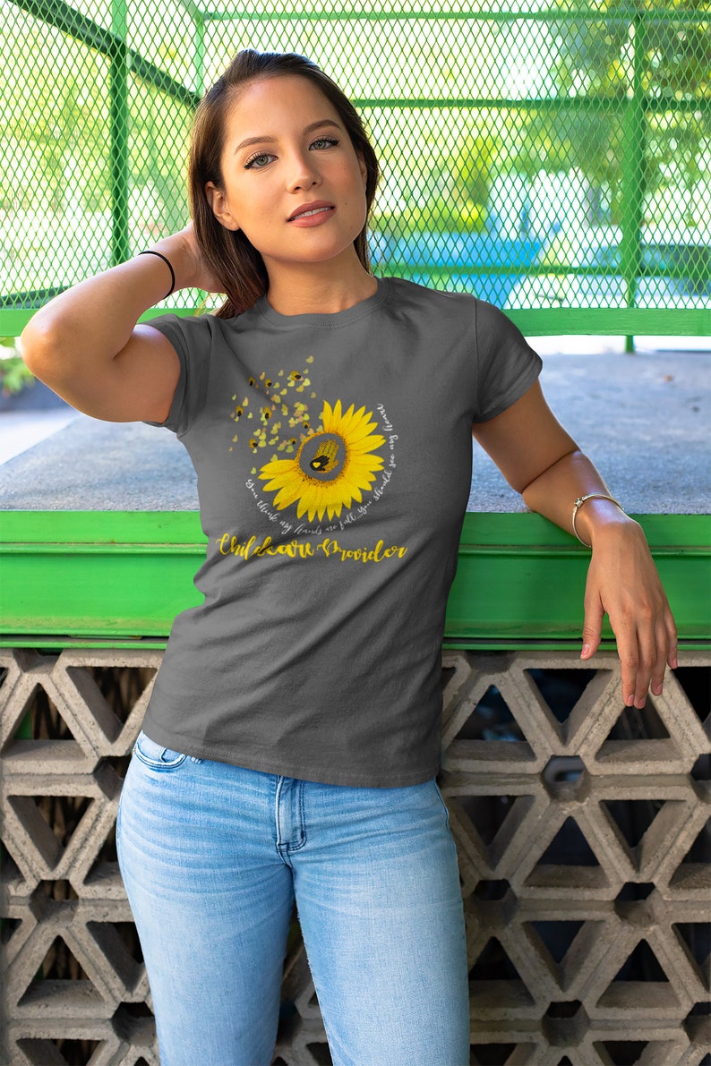 Woman wearing a gray t-shirt with a sunflower design and text, standing against a green metal fence.